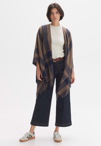 Brown and navy plaid poncho with fringes, featuring a loose fit. Paired with a white t-shirt, wide-leg dark denim pants, and sneakers.