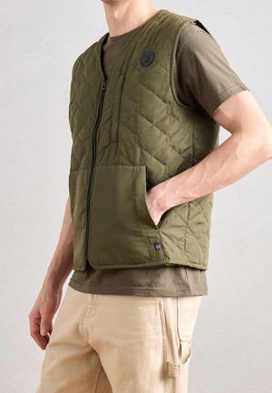 Homme portant une veste matelassée vert olive sur un t-shirt marron et un pantalon beige, avec une main dans la poche de la veste sur fond uni.