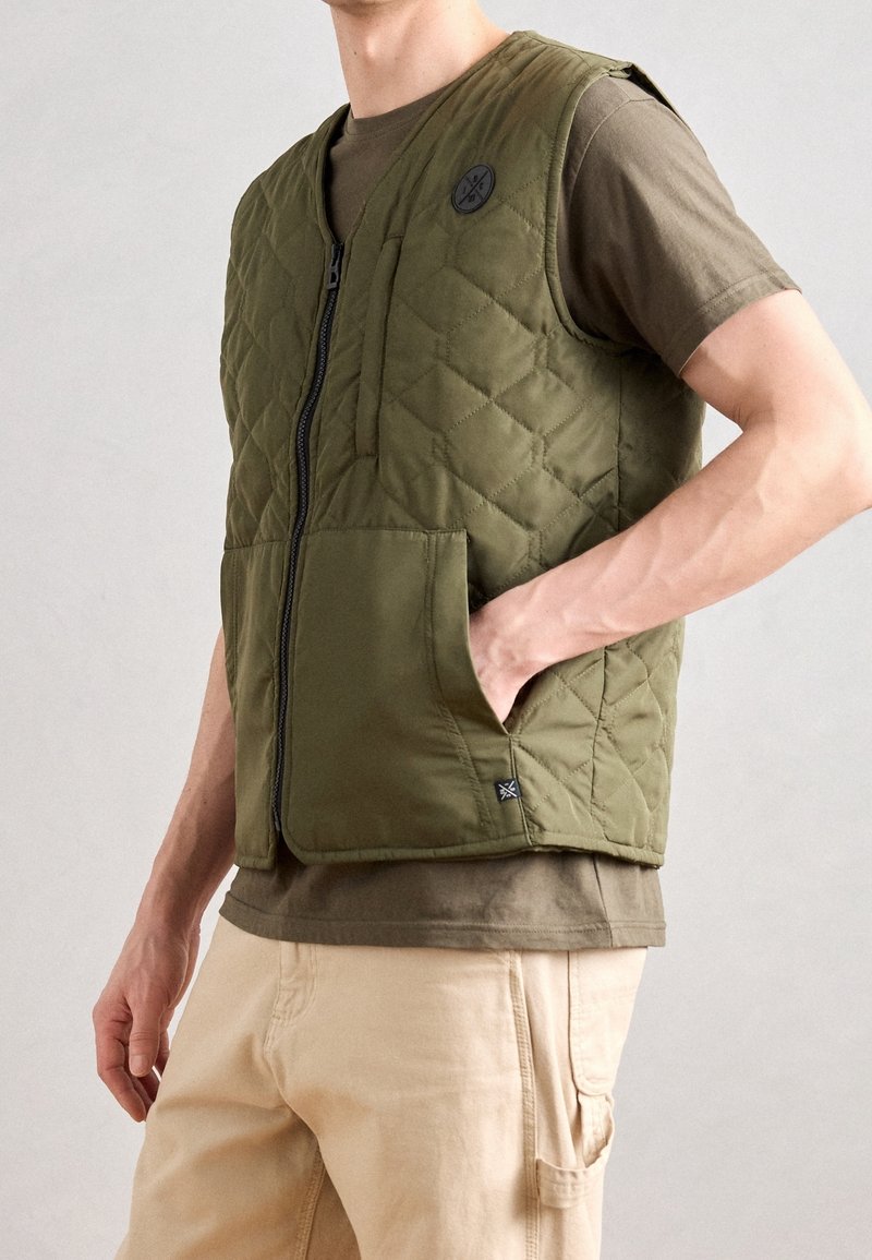 Homme portant une veste matelassée vert olive sur un t-shirt marron et un pantalon beige, avec une main dans la poche de la veste sur fond uni.