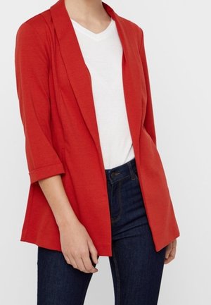 Femme portant un blazer rouge vif avec les manches retroussées sur un t-shirt blanc et un jean bleu foncé sur un fond uni.