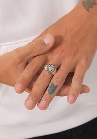 Sølvtone ring med et løvehoved sidder på en finger. Hænder viser forskellige hudtoner med synlige tatoveringer og små ufuldkommenheder. Baggrunden er neutral.