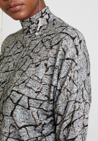 Suéter gris de cuello alto con un patrón texturizado y agrietado en negro y cobre. La tela parece suave y acogedora, con un ajuste holgado.