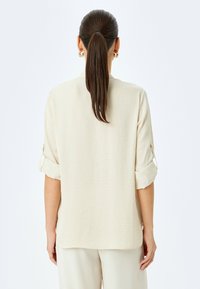Beżowa, lekka koszula z podwiniętymi rękawami, o teksturowanej tkaninie. Widok z tyłu pokazujący szew ramienia i linię dołu. Minimalistyczny design.