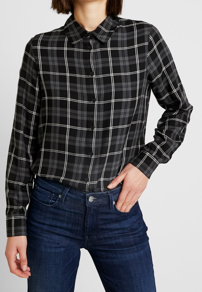 Femme portant une chemise à carreaux noire et grise boutonnée, rentrée dans un jean bleu foncé, avec une main dans une poche.
