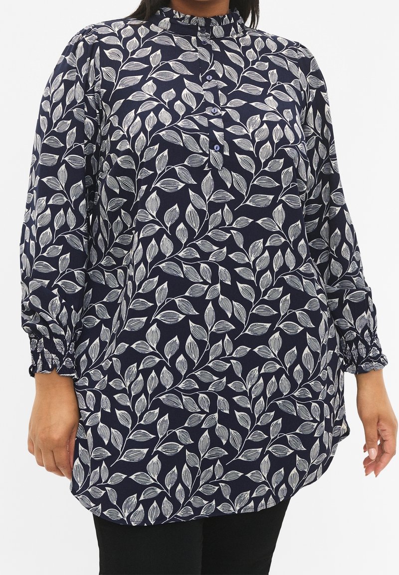 Femme portant un chemisier bleu marine à motif de feuilles blanches, boutonné à l'avant, col montant et poignets élastiques, associé à un pantalon noir.