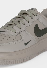 Ανοιχτό μπεζ αθλητικό παπούτσι Nike με λογότυπα Swoosh σκούρου πράσινου χρώματος, αντίστοιχα κορδόνια και υφή στη λαστιχένια σόλα σε κοντινή λήψη.