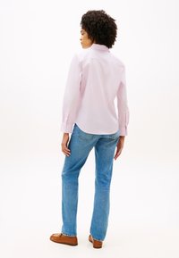 Γυναίκα με σγουρά μαλλιά στέκεται πλάτη, φοράει ανοιχτό ροζ πουκάμισο, μπλε τζιν και καφέ παπούτσια πάνω σε λευκό φόντο.