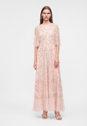 Frau steht in einem rosa transparenten bodenlangen Kleid mit Blumenstickerei und kurzen Ärmeln, kombiniert mit offenen High-Heels.