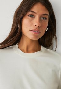 Sweatshirt de couleur crème avec un col rond et une texture douce. Le modèle a les cheveux longs et porte peu de bijoux, mettant en valeur sa coupe décontractée.