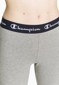 Szare legginsy z czarnym elastycznym paskiem w talii, z białym napisem "Champion", wykonane z miękkiego, elastycznego materiału.