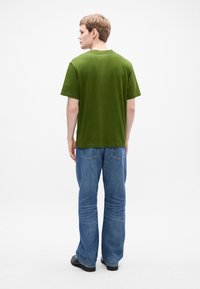Young man standing, facing away, wearing a green short-sleeve t-shirt, blue jeans, and black shoes against a white background.