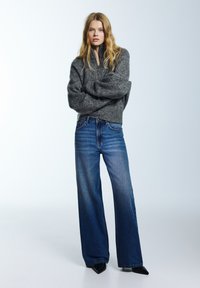 Szürke kötött pulóver magas nyakkal, széles szárú kék farmernadrággal párosítva. Világos háttér előtt, hegyes fekete cipőkkel.