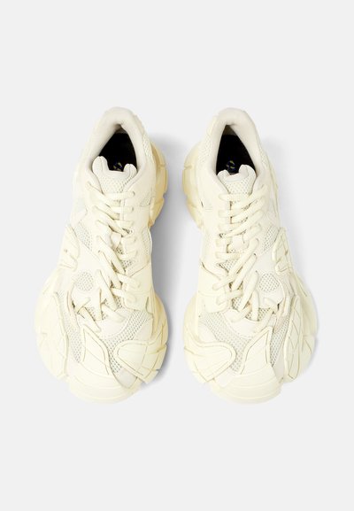 Cream-colored athletic shoes with a textured mesh upper, thick rubber sole, and distinct layered design. Features flat laces and padded tongue.