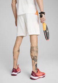 Beli športni šortsi, čevlji s rdečo-oranžno vzorčno zasnovo, pro paddel v roki, podrobne tetovaže na nogah, mrežasta majica z oranžnimi poudarki.