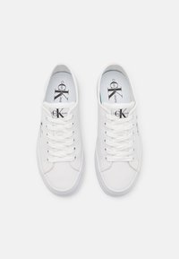 Weiße Segeltuch-Sneaker mit Schnürsenkeln, ausgestattet mit einer Gummisohle und einem bedruckten Logo auf der Zunge. Einfaches, klassisches Design mit runder Zehenpartie.