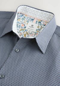Chemise à motifs bleus avec un extérieur texturé. Le col est bordé d'un tissu floral avec des accents roses et jaunes. Étiquette visible.