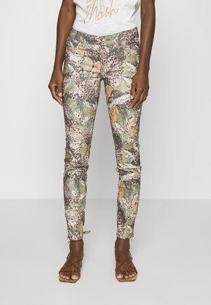 Spodnie o dopasowanym kroju z nadrukiem liści w kolorach zielonym, brązowym i beżowym oraz wzorem w panterkę, noszone z brązowymi sandałami na sznurowadła na ciemnoskórej osobie.