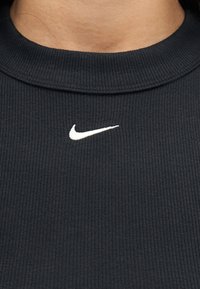 Μαύρο ριμπ κοντομάνικο με λαιμόκοψη και λευκό λογότυπο Nike Swoosh που είναι τοποθετημένο κεντρικά κάτω από το λαιμό στο πάνω μέρος του στήθους ενός ατόμου.