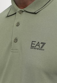 Green polo shirt with a textured fabric, three button placket, and black embroidered branding on the chest. Features contrasting collar trim.