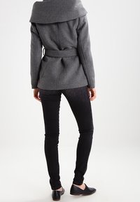 Abrigo de mezcla de lana gris con cinturón, cuello ancho, tejido texturizado y silueta ajustada, combinado con jeans negros ajustados y zapatos planos.
