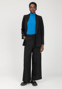 Blazer nero su misura sopra un dolcevita blu, abbinato a pantaloni neri a gamba larga e scarpe nere chunky. Texture morbide, design classico.