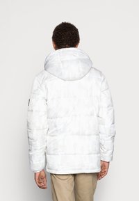 Uomo che indossa una giacca imbottita a motivo camouflage bianco con cappuccio, mostrato di spalle, in piedi contro uno sfondo chiaro e uniforme.