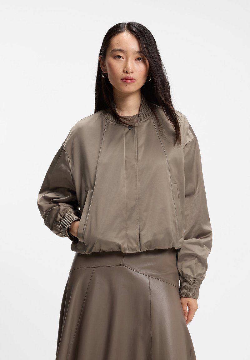 Femme aux longs cheveux foncés portant une veste bomber taupe avec des poignets côtes et une jupe en cuir assortie, debout devant un fond blanc.
