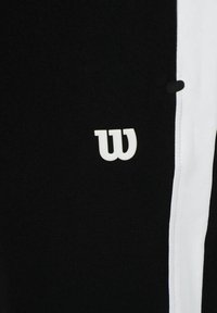 Zwarte sportbroeken met een witte zijstreep en een opvallend wit logo. De stof lijkt glad met een rekbare textuur.