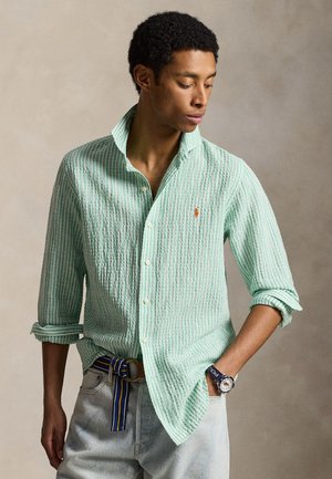 Homme portant une chemise à rayures vert clair avec les manches retroussées, un jean clair, une ceinture rayée et une montre-bracelet, regardant vers le bas sur le côté.