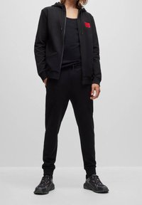 Persoon in een zwarte hoodie, zwarte tanktop, zwarte joggerbroek met rode patches en zwarte sneakers die tegen een effen grijze achtergrond staat.