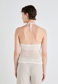 Heklet halterneck topp i kremfarge med et åpent vevmønster. Har en knyting i nakken og en tettsittende byste. Kombinert med beige stripete bukser.