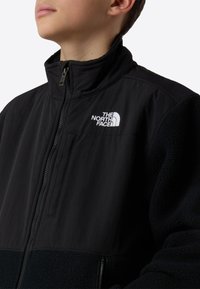 Crna jakna na patentni zatvarač s kombinacijom glatkog tkanina i mekane runaste tkanine, visokim okovratnikom, džepom na prsima i bijelim logotipom na lijevoj strani.