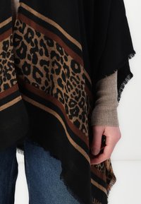 Poncho nero con un design a stampa leopardata, caratterizzato da strisce marroni e beige. I bordi sono frangiati. Indossato sopra un maglione grigio a costine e jeans blu.