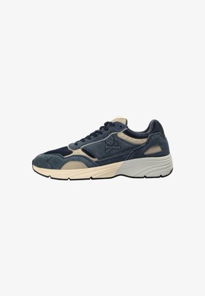 Zapatilla de ante y malla en azul marino y beige con cordones delanteros, suela blanca gruesa y logotipo de calavera con huesos cruzados en el lateral.