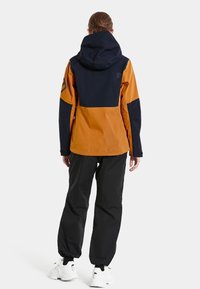 Waterproof jacket in orange and navy, featuring a hood, zippers, and elastic cuffs. Paired with black pants and white sneakers.