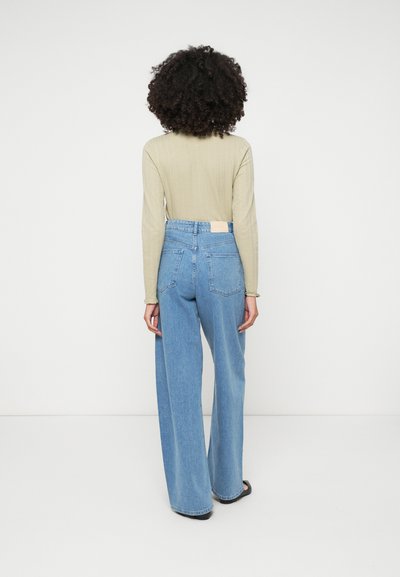 Svetlo zelena majica s dugim rukavima i visokim strukom, široke traperice plave boje s zadnjim džepovima i pravokutnom etiketom na struku.