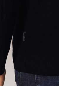 Schwarzer Strickpullover mit gerippter Struktur und einem seitlichen Etikett mit der Aufschrift "STATE of ART." Verfügt über eine figurbetonte Passform und elastische Bündchen.