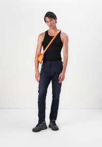 Čierny top na ramienka, námornícke džínsy a čierne tenisky. Modelka drží malú oranžovú crossbody tašku. Jednoduché biele pozadie.
