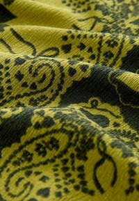 Zielony materiał o teksturowanej powierzchni, z ciemnym wzorem paisley oraz skomplikowanymi detalami widocznymi w fałdach i zagnieceniach.