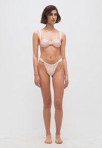 Žena so krátkymi tmavými vlasmi, oblečená v svetloružových bikinách s nariaseniami, stojaca bosá na čistom bielom pozadí.