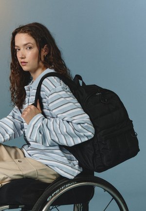 Mochila negra con correas acolchadas y cremalleras, llevada sobre una camisa de manga larga a rayas azul claro. Persona sentada en una silla de ruedas frente a un fondo azul.