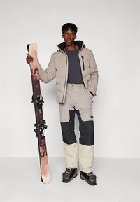 Suusavarustus, mis sisaldab beežikat polsterdatud jope, halli ja tumesinist suusapüksid ning musta värvi suusasaapaid. Käes hoitakse suuska, millel on tumedad kinnitused.