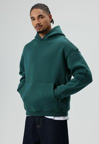 Donkergroene hoodie met capuchon, een voorzak, geribbelde boorden en zoom. Gedragen over een wit shirt, met een ontspannen pasvorm en een zachte textuur.