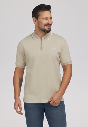 Uśmiechnięty mężczyzna z ciemnymi włosami i brodą, ubrany w beżową koszulkę polo z krótkim rękawem i zamkiem błyskawicznym oraz niebieskie dżinsy, stojący na jednolitym szarym tle.