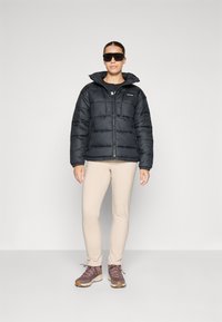 Zwarte gewatteerde jas met een hoge kraag, rits aan de voorkant, gedragen over een zwart shirt en lichtbeige aangesloten broek; gecombineerd met bruine wandelschoenen.