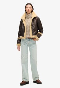 Chaqueta de cuero marrón con cuello y puños de piel de oveja beige, llevada sobre un suéter texturizado crema y jeans rectos azul claro. Zapatos marrones.