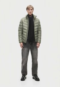 Ανοιχτό πράσινο επενδυμένο μπουφάν με κάθετο ραμμένο σχέδιο, μαύρο κασκόλ, σκούρα τζιν και μαύρα παπούτσια. Το μοντέλο στέκεται απέναντι από ένα ουδέτερο φόντο.