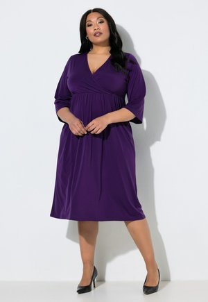 Donna che indossa un vestito viola al ginocchio con maniche a tre quarti e tacchi neri, in piedi su sfondo bianco.