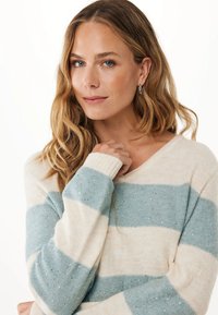 Frau mit welligem, blonden Haar, die silberne Ohrhänger trägt und einen cremefarbenen und hellblauen gestreiften Pullover mit glitzernden Details trägt, vor einem weißen Hintergrund.