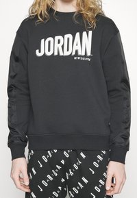 Sudadera negra con "JORDAN" en letras blancas en negrita. Tiene mangas texturizadas y un corte holgado. Combinada con pantalones negros que presentan un logo repetido.